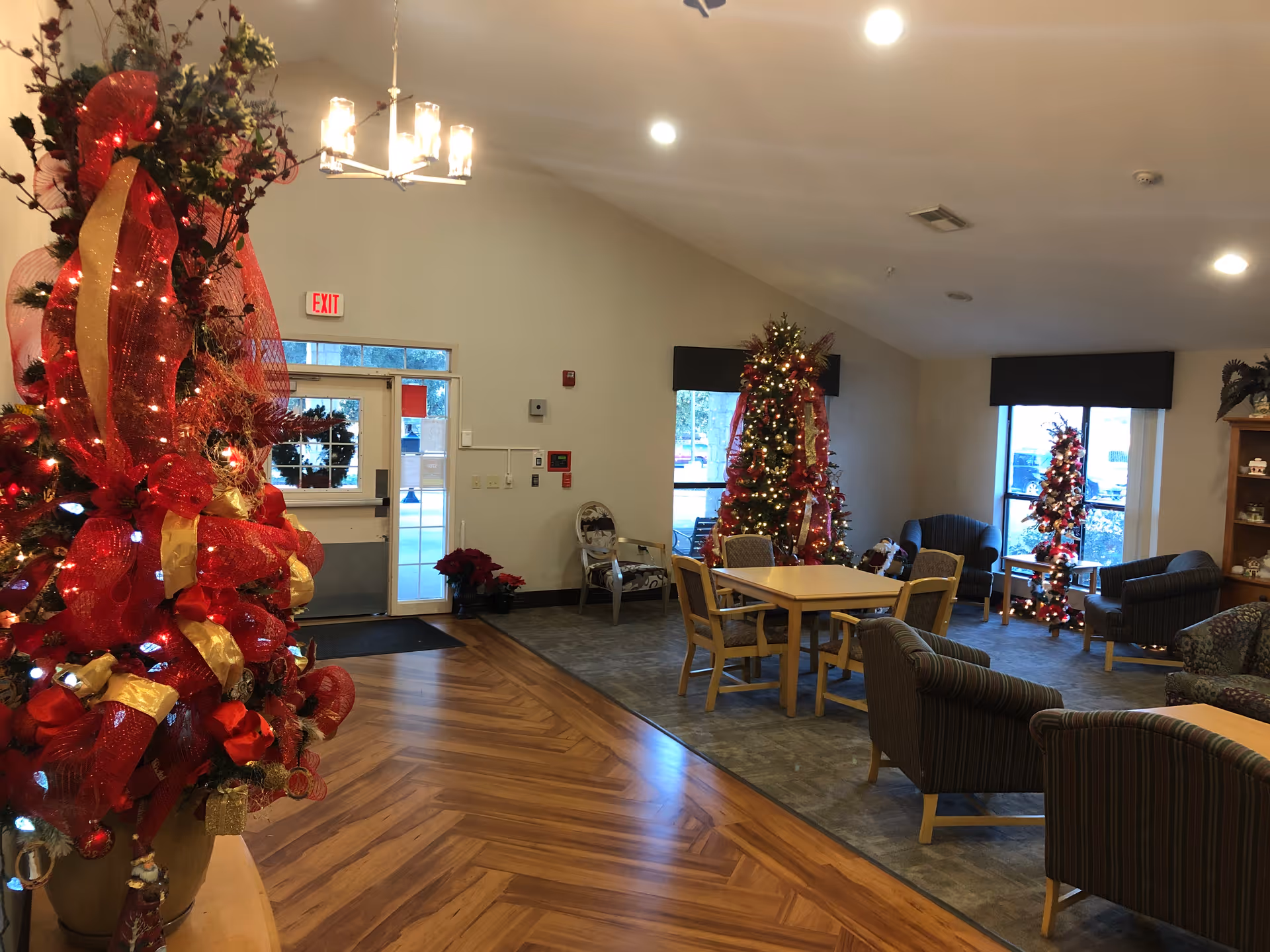 A cozy common area decorated for Christmas with multiple Christmas trees adorned with red and gold ribbons and lights. The room has wooden flooring and carpeted seating areas with chairs and tables. Large windows let in natural light, and there is an exit door with a wreath on it.