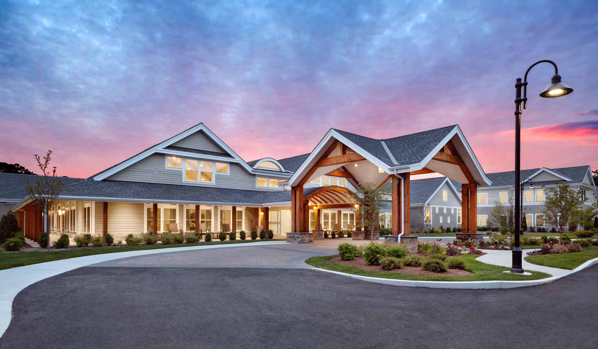 Exterior view of a senior living facility named Maplewood at Brewster during sunset, showing a large building with a covered entrance supported by wooden beams, well-maintained landscaping, and a street lamp on the right side.