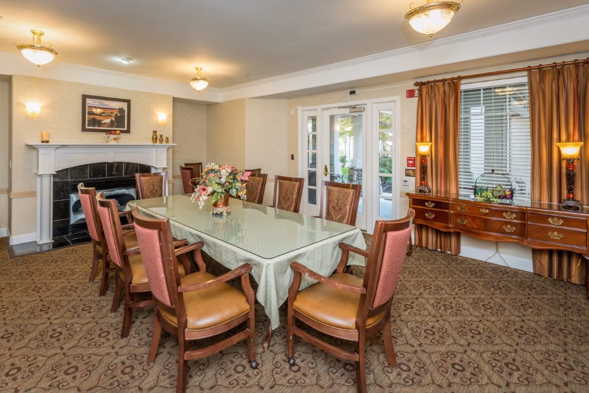 Formal dining room with a long glass-topped table surrounded by upholstered chairs, a floral centerpiece, fireplace, and sideboard by curtained windows.