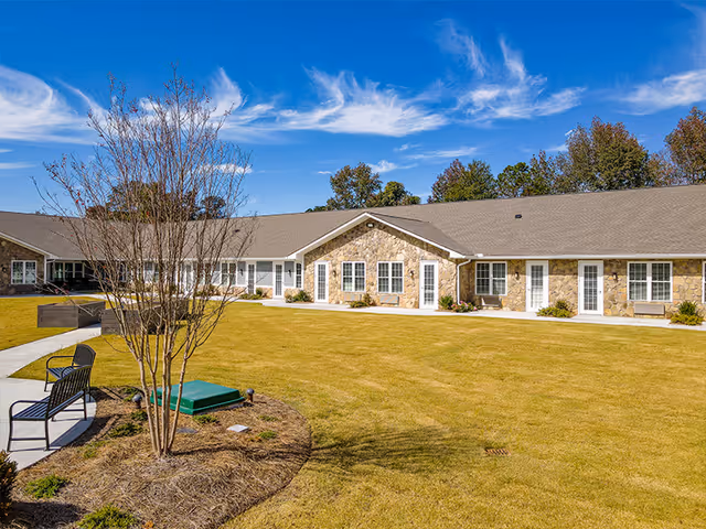 Single-story assisted living facility building with stone facade and multiple white doors and windows, surrounded by a well-maintained lawn and a few trees under a blue sky with wispy clouds. There are benches and a paved walkway in the foreground.