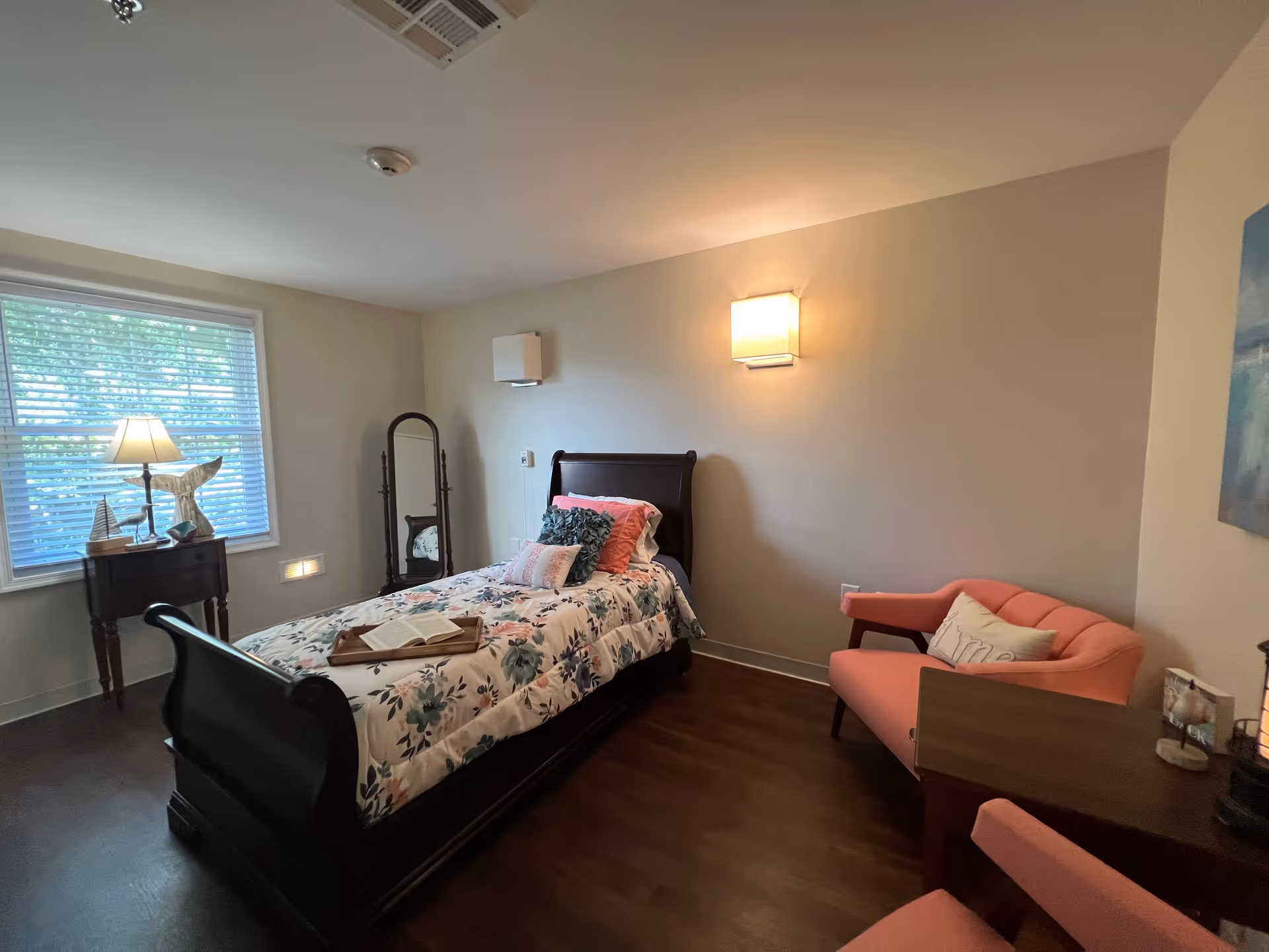 A cozy bedroom with a single dark wooden sleigh bed adorned with floral bedding and multiple pillows. There is a small wooden side table with a lamp and decorative items next to a window with blinds. A full-length mirror stands against the wall. On the right side, there is a pink upholstered loveseat with a pillow, and a wooden desk with a matching pink chair. The room has wooden flooring and neutral-colored walls with a wall-mounted light fixture.