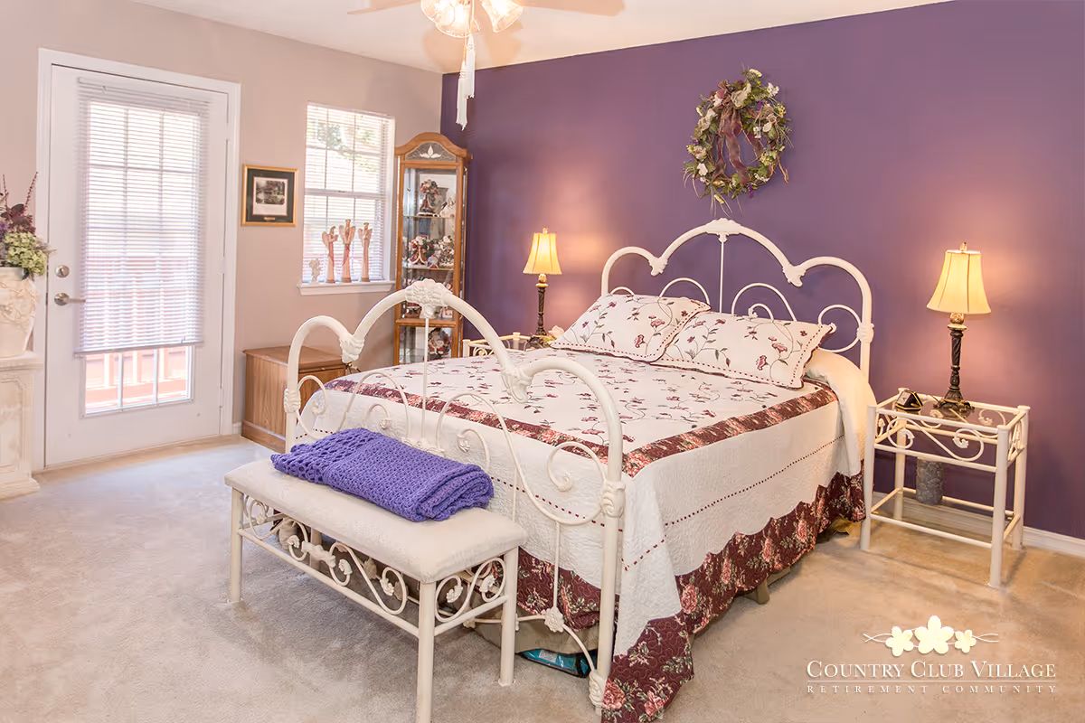 A cozy bedroom with a white metal bed frame and floral bedding. The wall behind the bed is painted purple and decorated with a floral wreath. There are two bedside tables with lamps on each side of the bed. A cushioned bench with a purple blanket is placed at the foot of the bed. The room has a window and a glass door with blinds, allowing natural light to enter. A wooden cabinet with decorative items is positioned near the window.