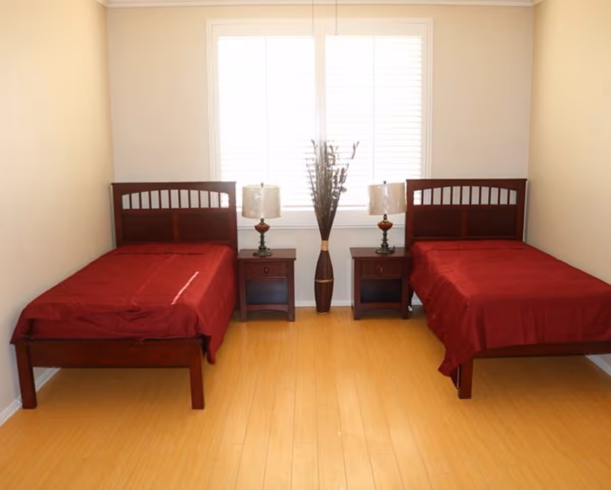A simple bedroom with two single beds featuring dark wooden frames and red bedspreads. Between the beds are two matching nightstands, each with a lamp. A tall vase with decorative branches is placed in the center under a window with white blinds. The room has light-colored walls and a wooden floor.