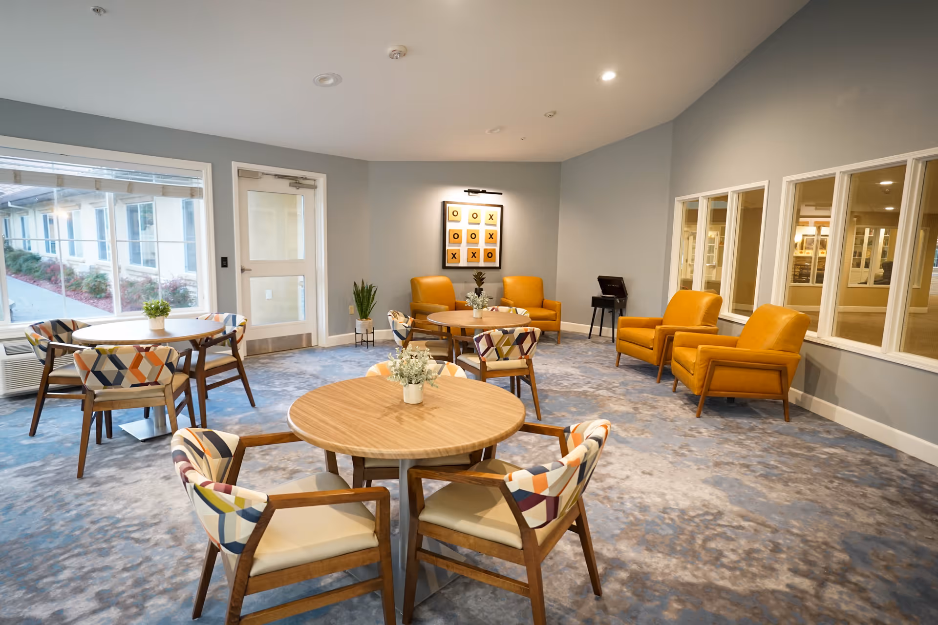 A bright and spacious common area with round wooden tables surrounded by colorful patterned chairs. There are three mustard yellow armchairs arranged against the wall, with a framed tic-tac-toe artwork above them. Large windows allow natural light to enter, and there are small potted plants on the tables and floor.