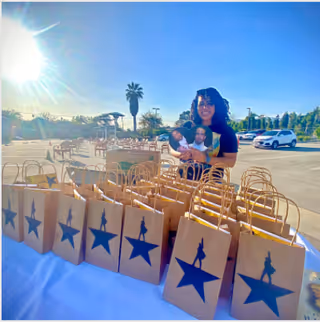 A woman standing behind a table filled with brown paper gift bags featuring a blue star and silhouette design, outdoors in a sunny parking lot with palm trees and cars in the background.