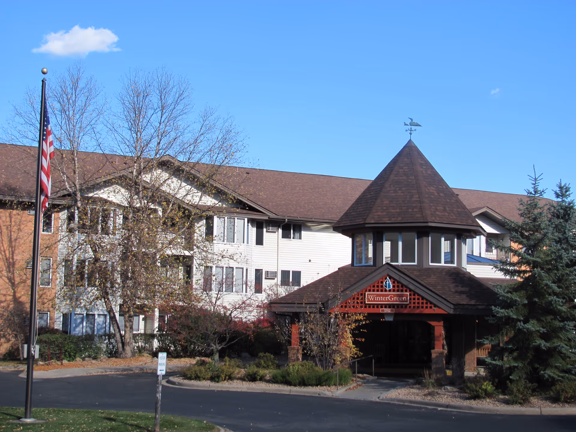 Exterior view of Wintergreen Independent Senior Living facility showing a multi-story building with a peaked roof entrance, surrounded by trees and landscaping under a clear blue sky. An American flag is visible on a flagpole in front of the building.
