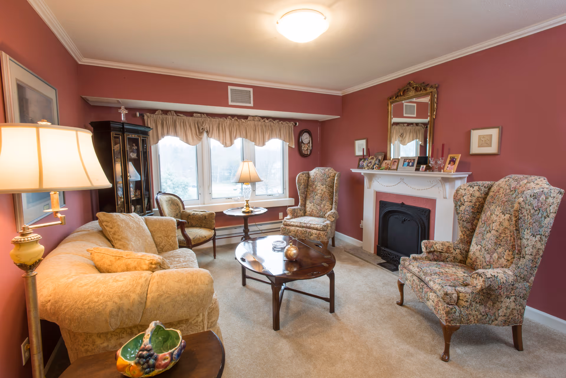 Cozy residential living room with floral wingback chairs, a beige sofa, coffee table, fireplace mantel, and a large bay window.