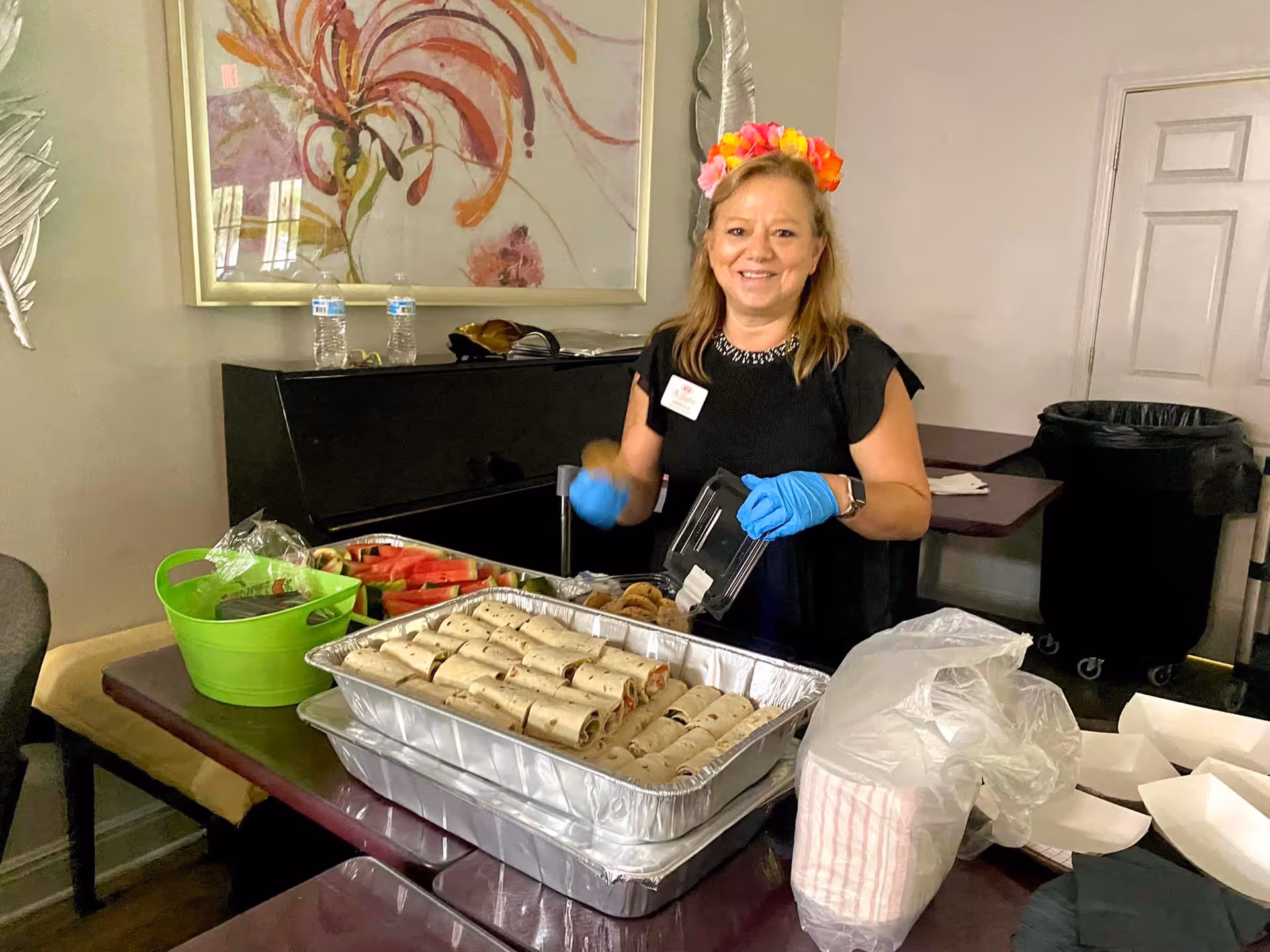 A smiling woman wearing a flower headband and blue gloves stands behind a table with trays of rolled sandwiches, watermelon slices, and cookies in a room with a piano and abstract wall art.