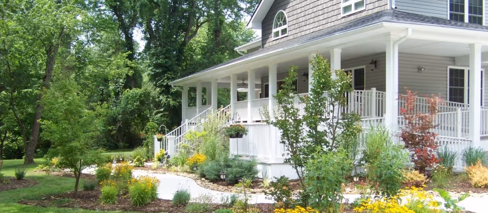 Front exterior of a light-gray assisted living building with a wraparound white porch surrounded by landscaped gardens and trees.