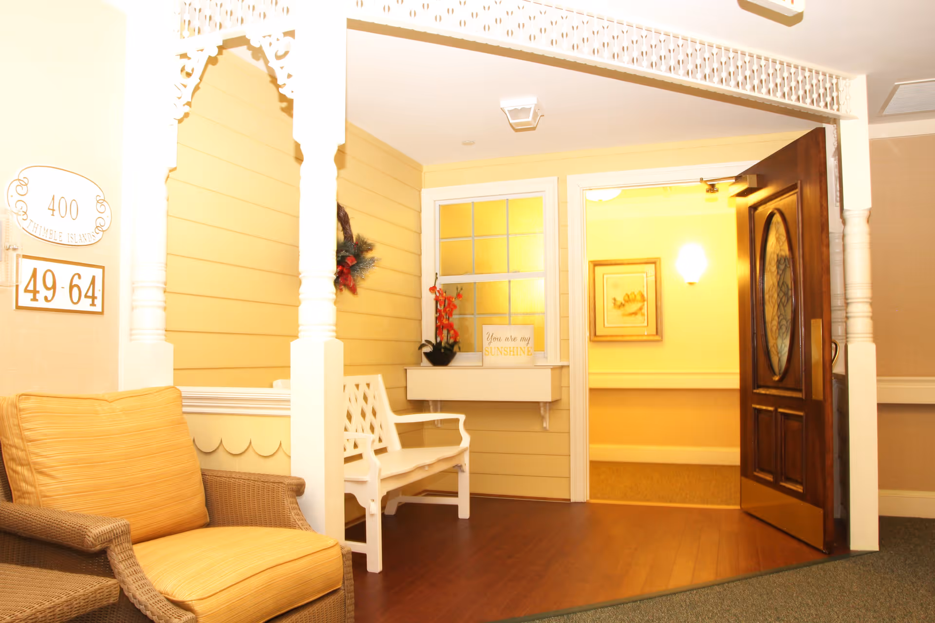 A cozy indoor seating area with a wicker chair featuring a yellow cushion and a white wooden bench against a yellow paneled wall. There is a decorative archway with intricate white trim, a small shelf with a potted plant and a sign that reads 'You are my SUNSHINE,' and an open wooden door leading to a warmly lit hallway with framed artwork on the wall.
