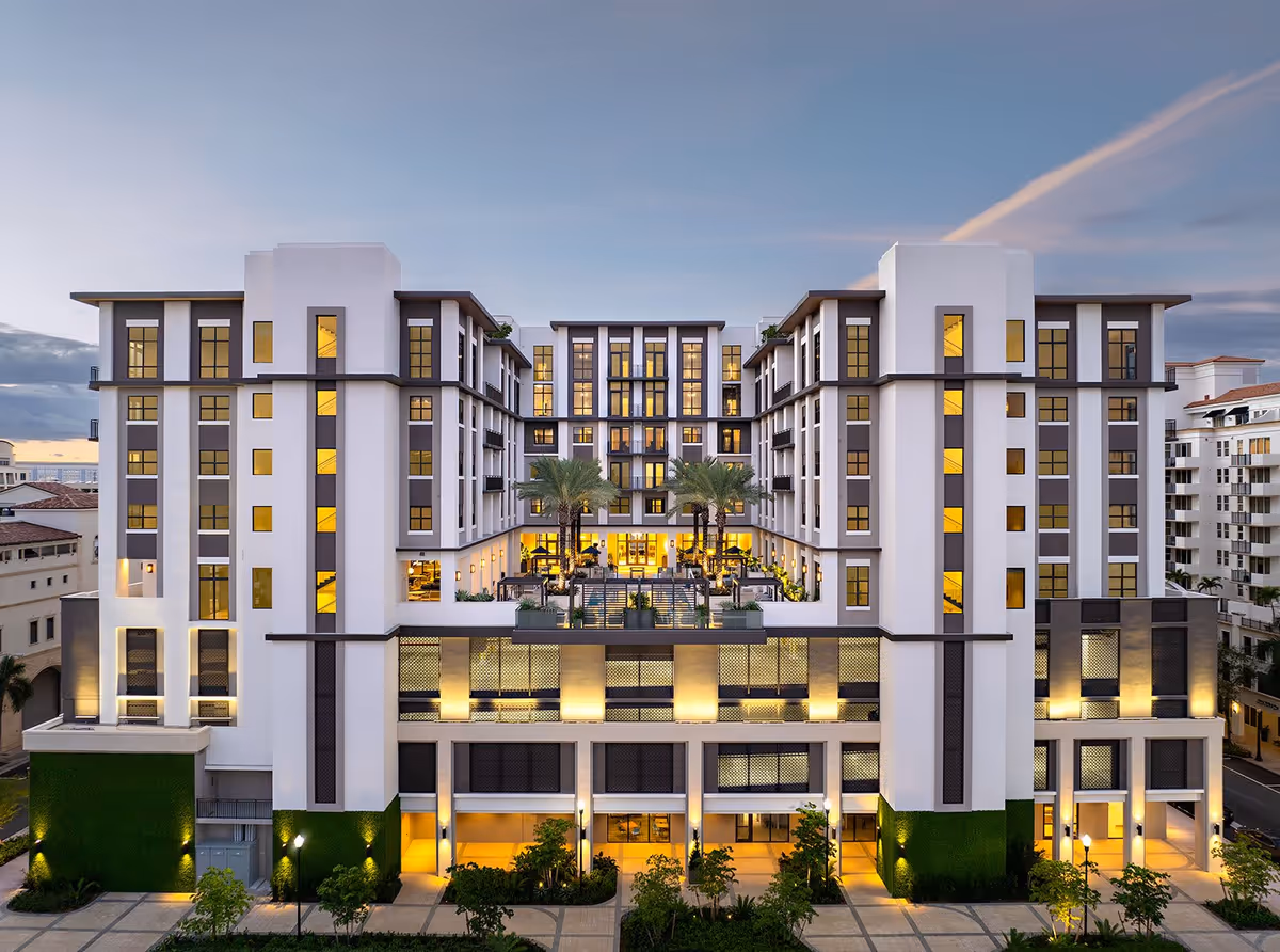 Exterior view of Belmont Village Senior Living Coral Gables building at dusk, showing a modern multi-story structure with illuminated windows and a central courtyard featuring palm trees and outdoor seating.