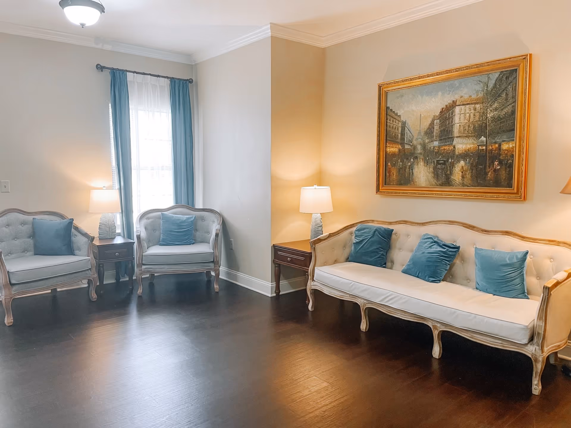 A cozy living room with a wooden floor, featuring a beige sofa with three blue cushions, two matching armchairs with blue cushions, two side tables with lamps, a window with blue curtains, and a large framed painting of a city street scene on the wall.