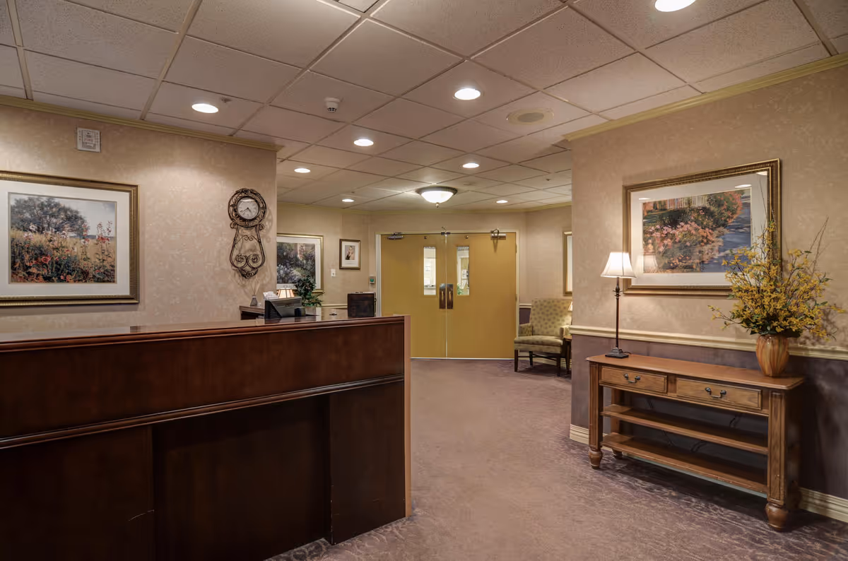 Reception area of a skilled nursing and rehabilitation center with a wooden front desk, beige walls, framed floral artwork, a decorative wall clock, a wooden console table with a lamp and a vase of yellow flowers, and double doors in the background.