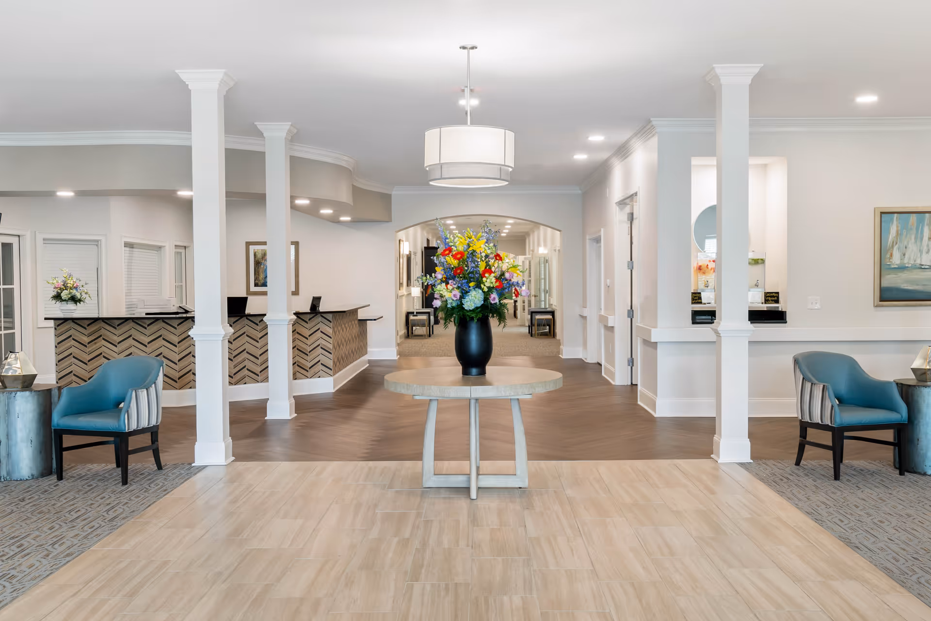 Bright senior living lobby with a central round table holding a large floral arrangement, reception desk and seating.