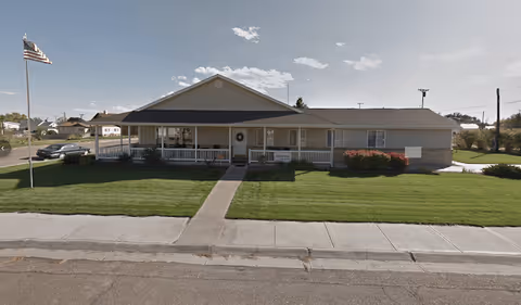 Single-story building with a covered front porch, beige siding, and a well-maintained lawn. An American flag is flying on a flagpole to the left of the building. The sky is clear with a few clouds.
