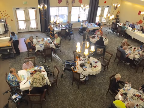 A dining room in an assisted living facility with several elderly residents seated at round tables. The tables are set with tablecloths, menus, and some food items. The room has large windows letting in natural light and decorative wall accents. Some residents are in wheelchairs, and staff members are assisting them.