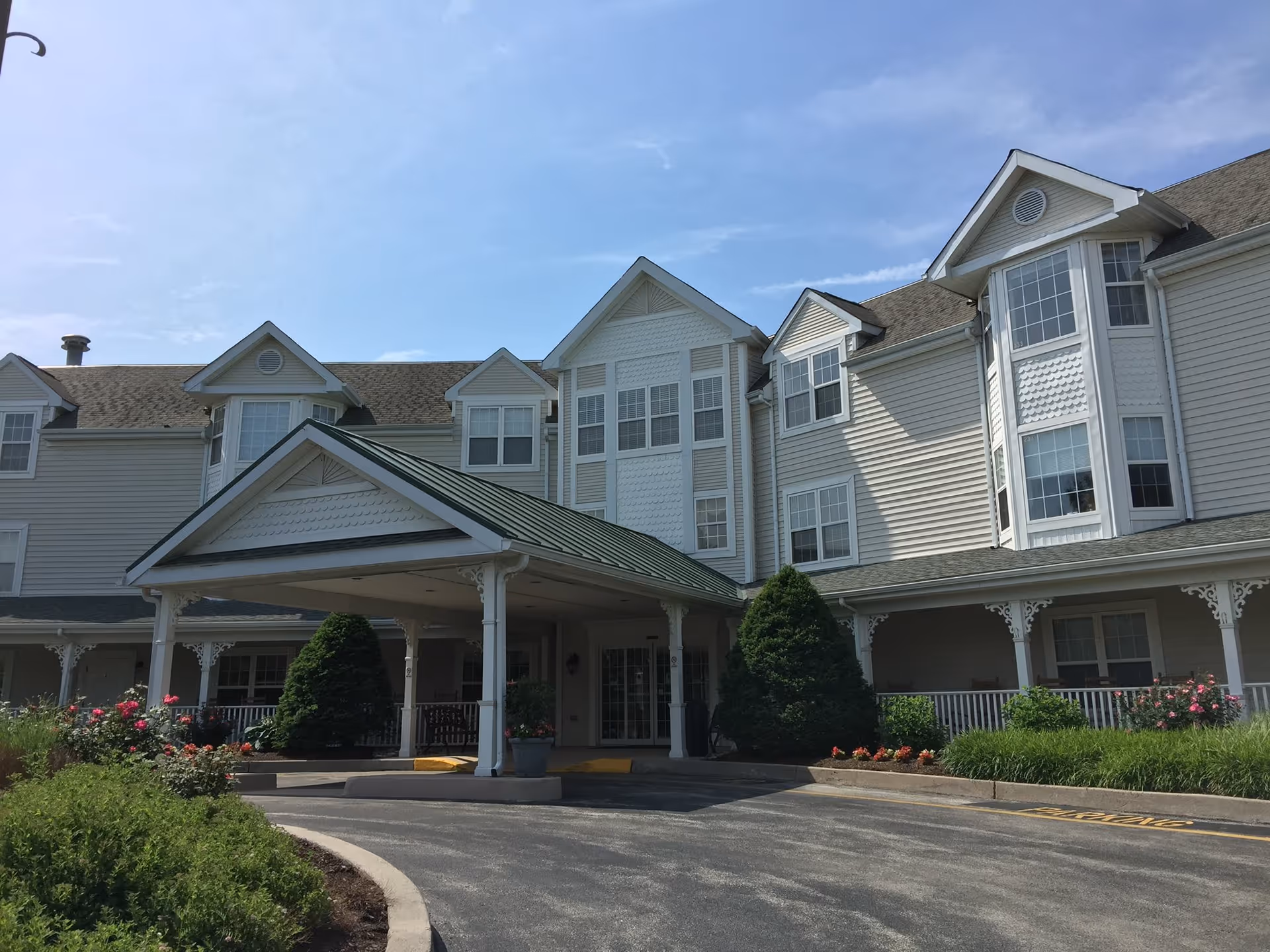 Front entrance of a multi-story senior living building with a covered porte-cochere, bay windows, and landscaped flower beds.