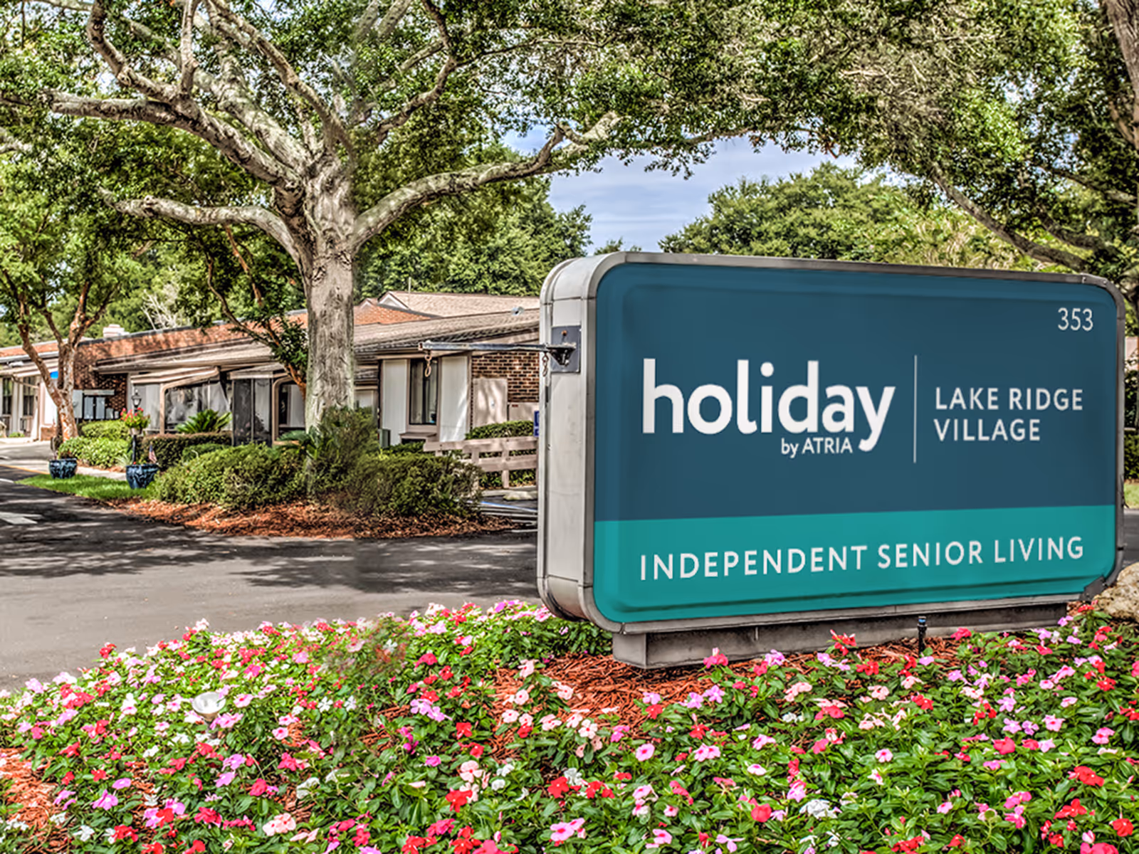 Entrance sign for Holiday Lake Ridge Village reading 'Independent Senior Living' by a flowerbed with low-rise buildings and large trees in the background.