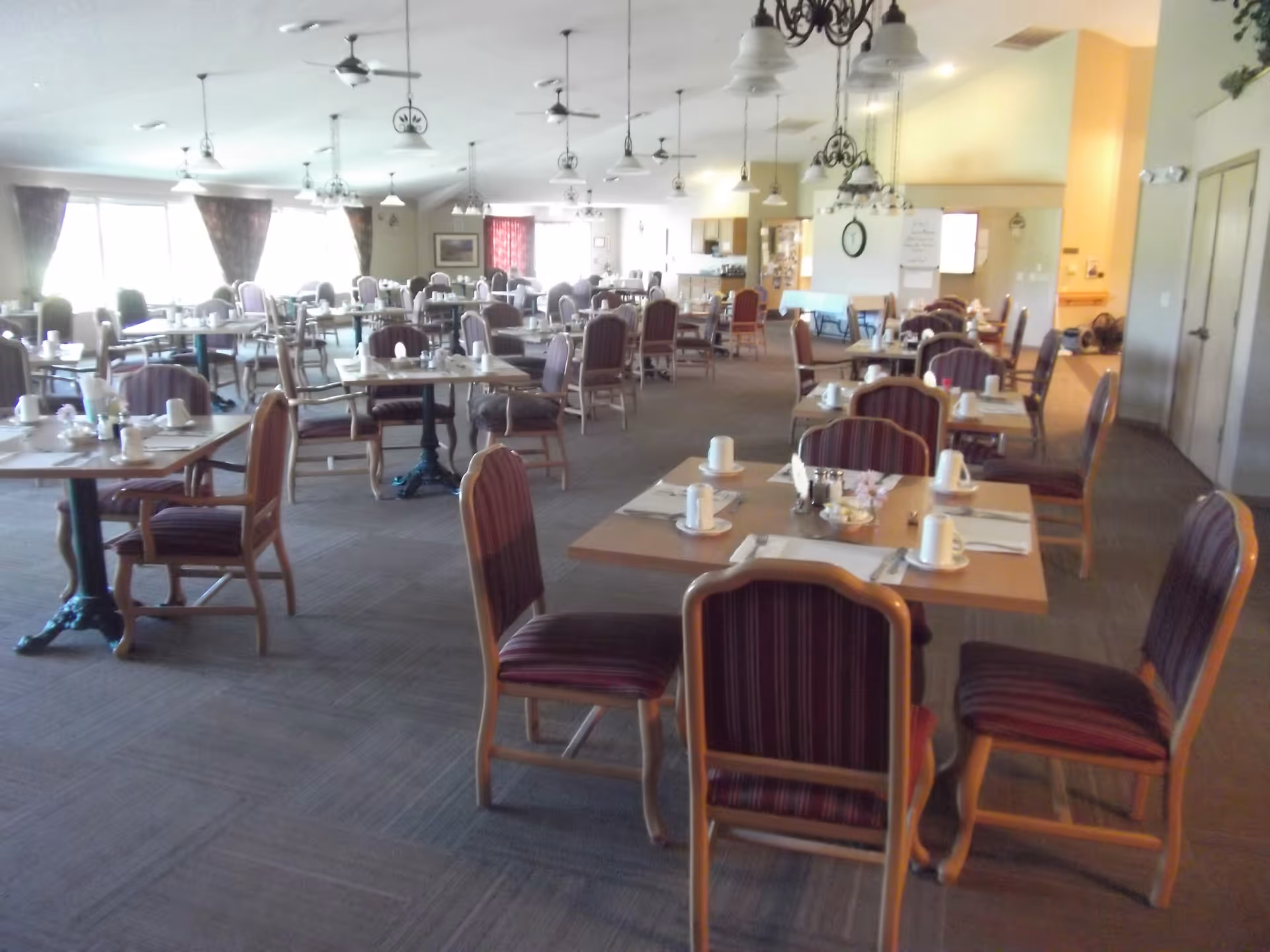 A spacious dining room with multiple tables and chairs arranged neatly. Each table is set with white cups, plates, and silverware. The room has large windows with curtains allowing natural light to enter, ceiling fans, and hanging light fixtures. The overall ambiance is bright and welcoming.