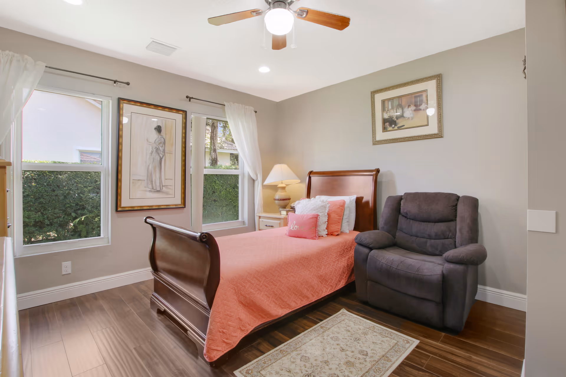 A cozy bedroom with a wooden sleigh bed covered with a coral quilt and multiple pillows. Next to the bed is a white nightstand with a lamp. A comfortable dark gray recliner chair is positioned beside the bed. The room has two windows with white curtains, framed artwork on the walls, a ceiling fan with a light, and a small area rug on the wooden floor.