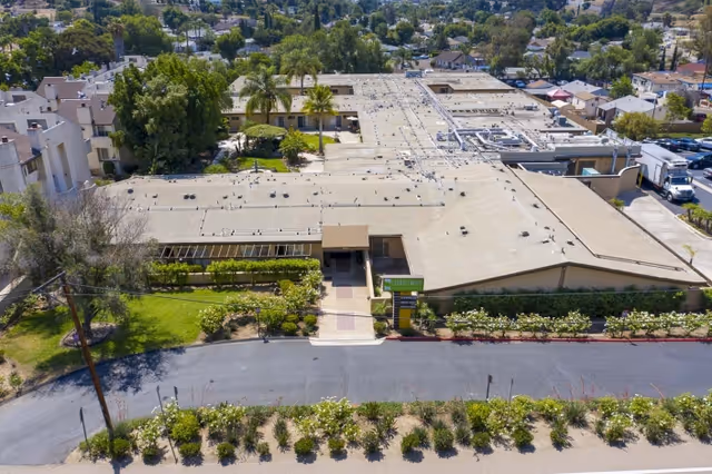 Aerial view of Lemon Grove Care And Rehabilitation Center showing a large, single-story building with a flat roof surrounded by greenery, trees, and a parking area. The entrance is visible with a sign and a pathway leading to the door.