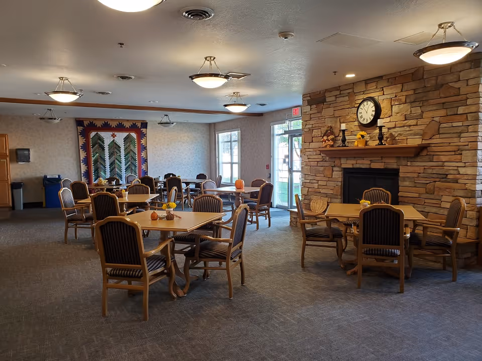 A dining room in a senior living facility with several wooden tables and chairs arranged neatly. The room features a stone fireplace with a clock and decorative items on the mantel. There are ceiling lights, a quilt hanging on the far wall, and large windows and a glass door letting in natural light.