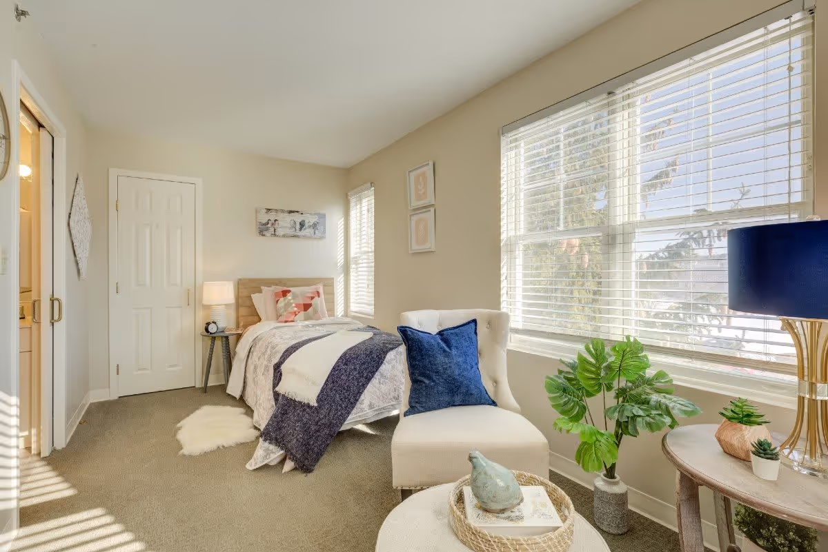 A bright bedroom with a single bed covered in a white and blue blanket, a small side table with a lamp and alarm clock, a white cushioned chair with a blue pillow, a round table with decorative plants and a blue lamp, and large windows with white blinds letting in natural light.