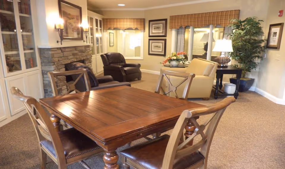 A cozy living room area with a wooden table and four chairs in the foreground. In the background, there are two leather armchairs, a beige armchair, a side table with a lamp, framed pictures on the walls, a large plant, and built-in white cabinets with glass doors. The room has warm lighting and a carpeted floor.