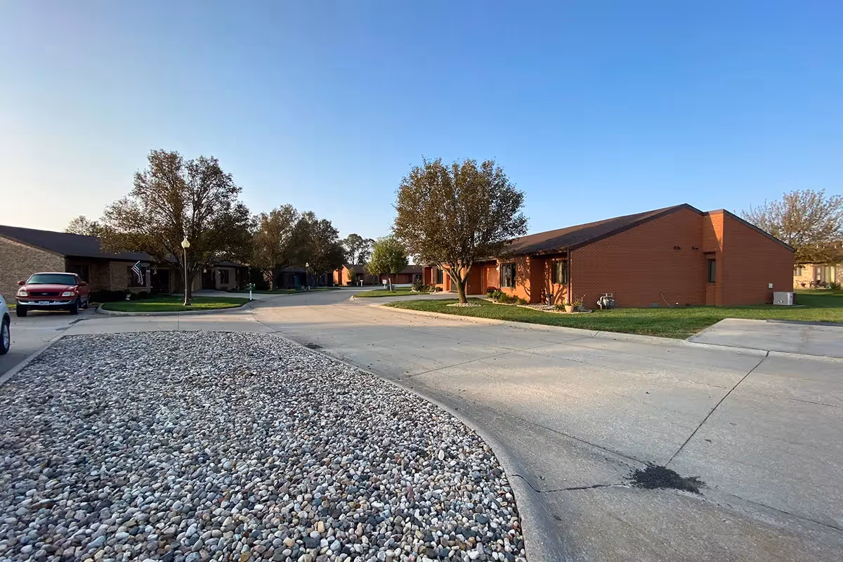 Exterior view of a single-story brick senior living campus with trees, parked cars, and a rocky median along a curved street.