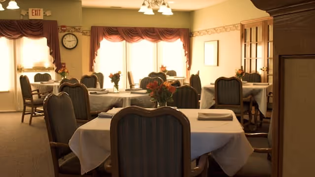 A warmly lit dining room with several tables covered in white tablecloths, each set with napkins and small floral centerpieces. The room has upholstered chairs around the tables, large windows with curtains allowing natural light, and a clock on the wall near an exit door.