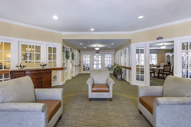 Bright communal sitting area with three armchairs facing a corridor flanked by glass‑paneled rooms and a reception desk.