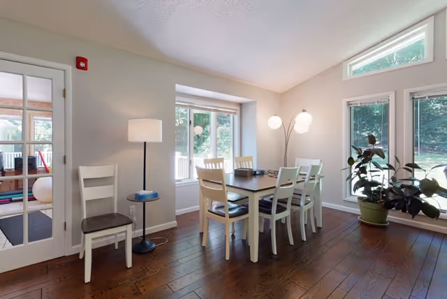A bright dining area with a rectangular wooden table surrounded by six white chairs with dark cushions. The room has large windows letting in natural light and a view of greenery outside. There is a floor lamp with three spherical lights near the corner and a potted plant by the window. A white chair and a standing lamp are positioned near a glass door leading to another room.