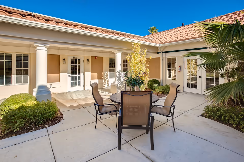 Outdoor courtyard area with a round table and four chairs surrounded by plants and shrubs, adjacent to a building with beige walls, white columns, and multiple glass doors under a tiled roof, under a clear blue sky.