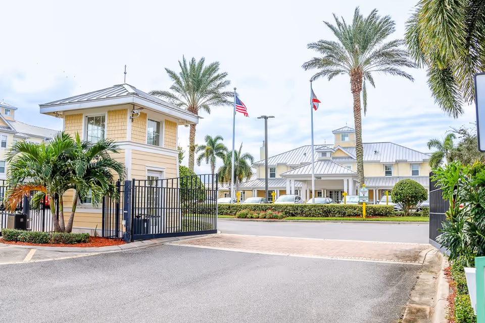Entrance gate and guardhouse of The Brennity at Melbourne Senior Living facility with palm trees, American and Florida state flags, and a large building in the background under a cloudy sky.