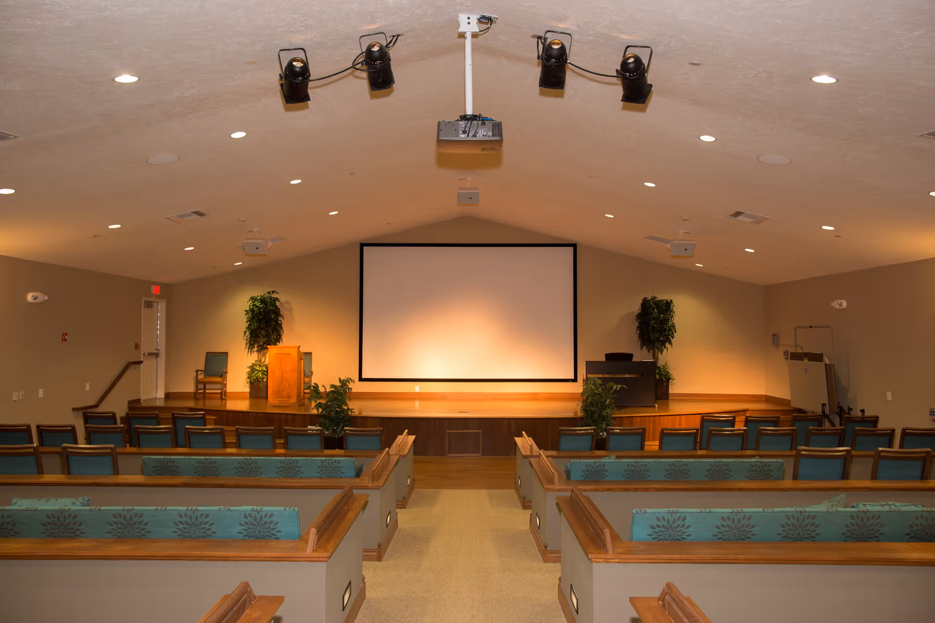 Interior view of a presentation or meeting room with rows of wooden pew-style seating with teal cushions facing a stage. The stage has a wooden podium, two potted plants, a chair, and a large blank projection screen on the back wall. Ceiling lights and mounted spotlights illuminate the room.