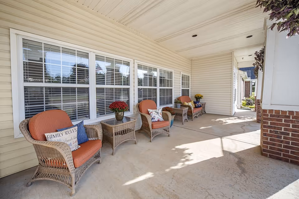 A covered outdoor patio area with four wicker chairs featuring orange cushions and decorative pillows, two wicker side tables with potted flowers, and large windows with white blinds on the building wall.