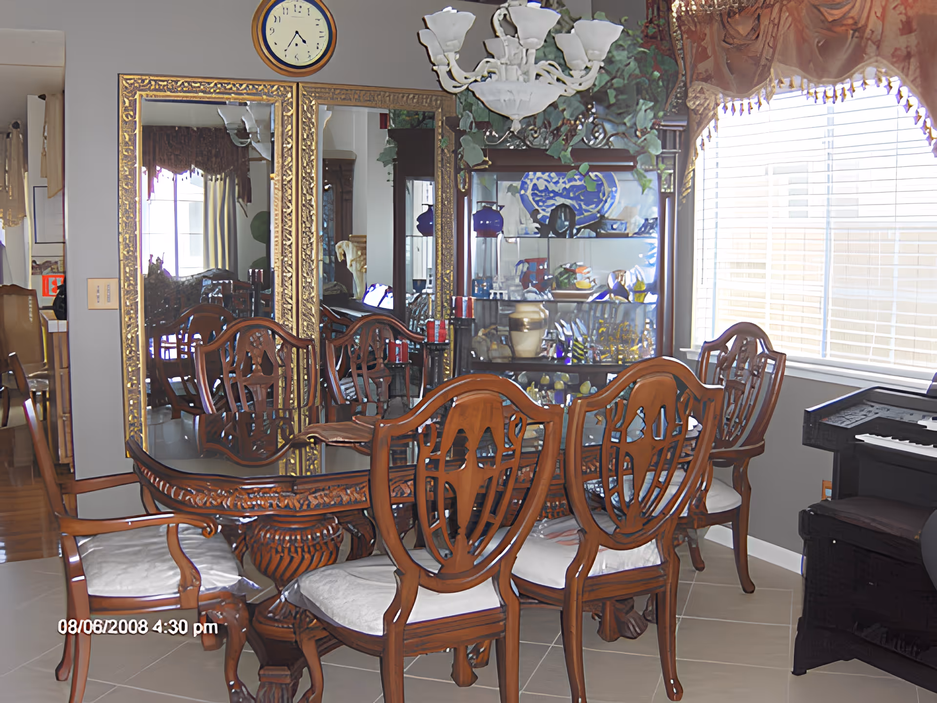 A dining room with a wooden dining table and six matching chairs with white cushions. Behind the table is a large mirror with a gold ornate frame and a clock above it. To the right, there is a glass cabinet displaying various decorative items and a window with blinds and a valance. A chandelier hangs above the table, and a piano is partially visible on the right side.