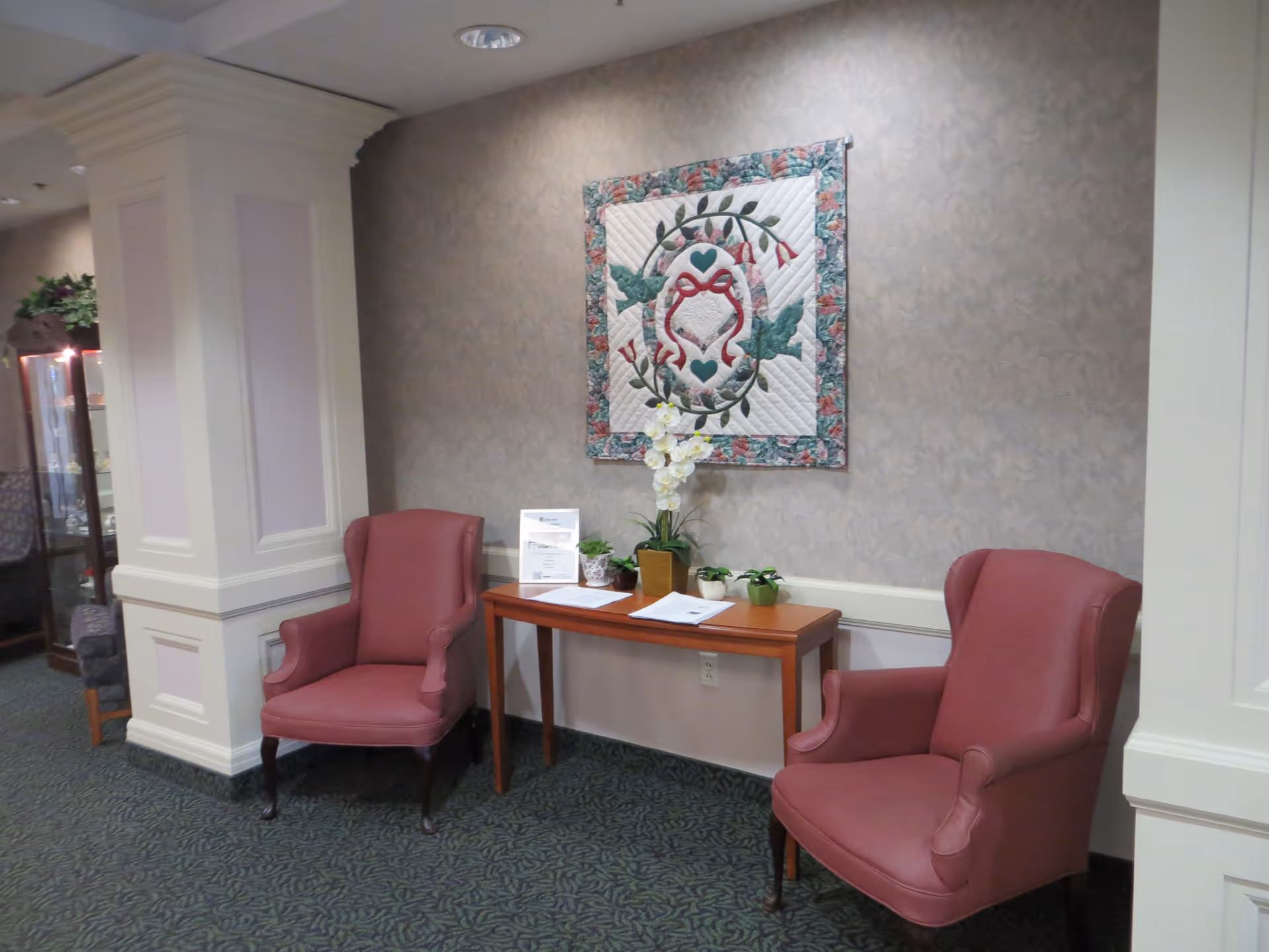 A cozy seating area in a senior living facility with two pink upholstered armchairs placed on either side of a wooden console table. The table holds a white orchid plant, small potted plants, and some papers. A decorative quilt with a floral and heart design hangs on the wall above the table. The area has patterned carpet and light-colored wallpaper with a subtle floral pattern.