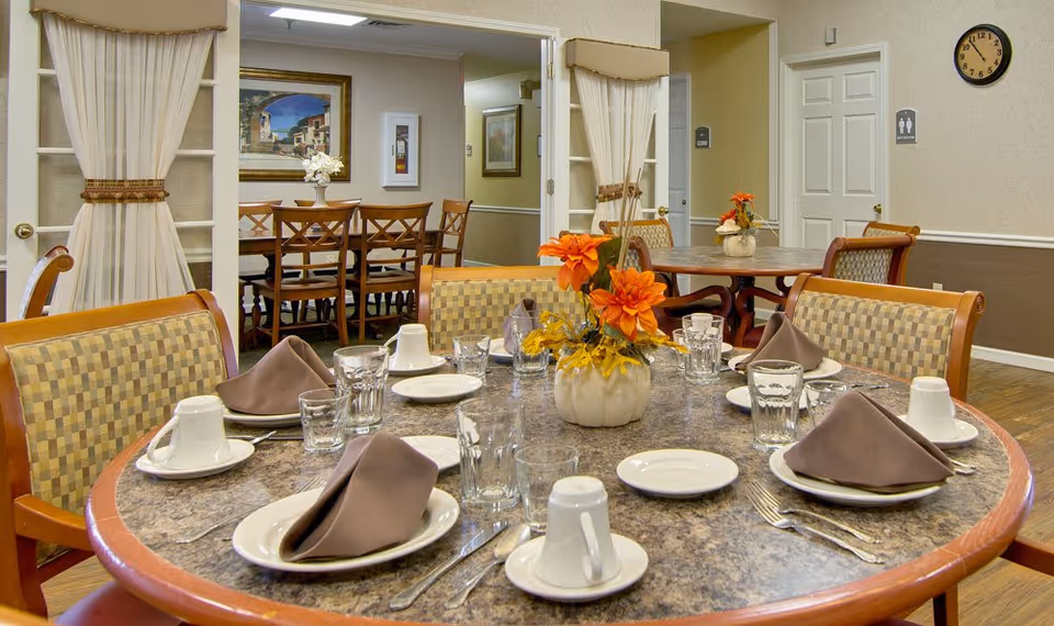 A dining room in a senior living facility with round tables set for a meal. Each table has plates, cups, glasses, silverware, and brown folded napkins. There are floral centerpieces with orange flowers on the tables. The room has beige walls with framed artwork and a clock on the wall. There are wooden chairs with patterned cushions around the tables.