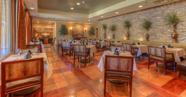 A spacious dining room with multiple tables covered in white tablecloths, each set with folded napkins and glassware. The room features warm lighting, a stone accent wall with decorative plants, and large windows with curtains allowing natural light to enter.