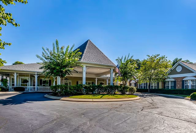 Exterior view of Rock Hill Grove Senior Living facility showing a covered entrance with a peaked roof, surrounded by trees and shrubs under a clear blue sky.