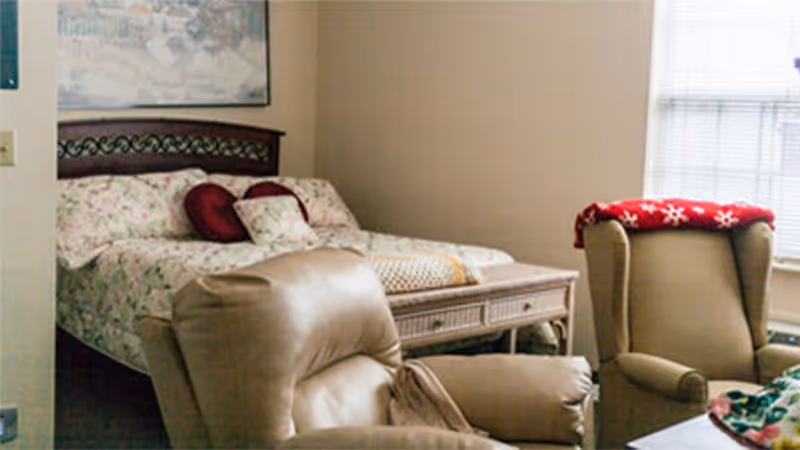 A cozy bedroom in an assisted living facility featuring a bed with floral bedding and red accent pillows, a wooden headboard, a wicker bench at the foot of the bed, and two beige recliner chairs, one with a red blanket with white snowflake patterns draped over it, near a window with blinds.