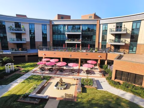 Outdoor courtyard area of a senior living facility with a central fountain, surrounded by benches and tables with pink umbrellas. The courtyard is enclosed by a multi-story building with balconies and large windows.