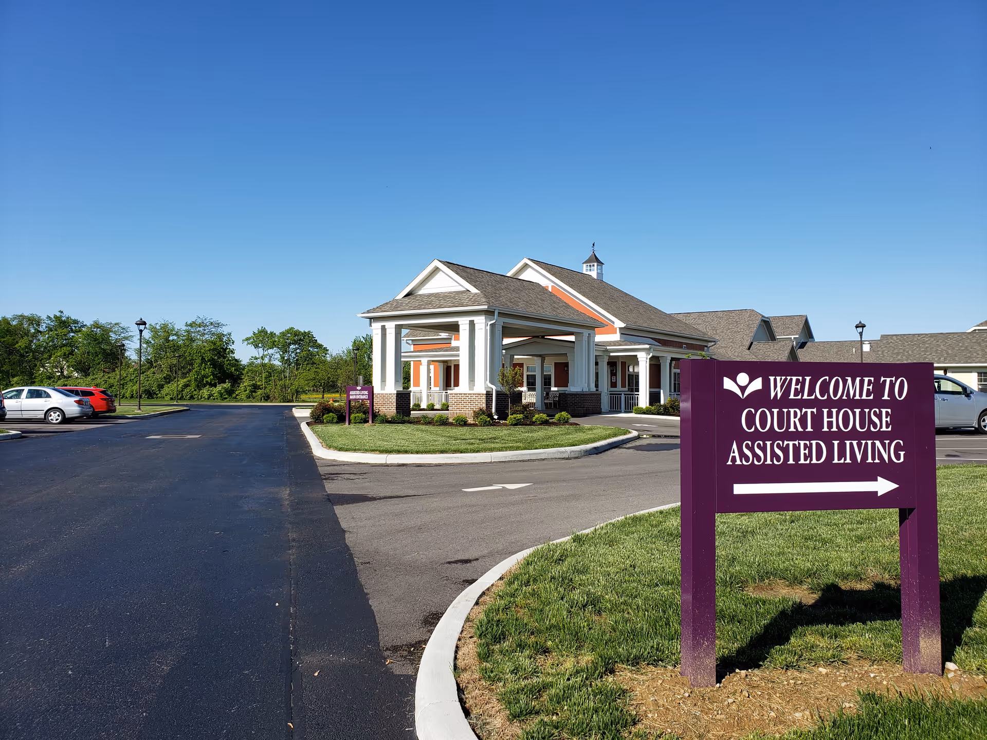 Exterior front view of an assisted living building, parking lot, and a purple sign reading 'WELCOME TO COURT HOUSE ASSISTED LIVING' under a clear blue sky.