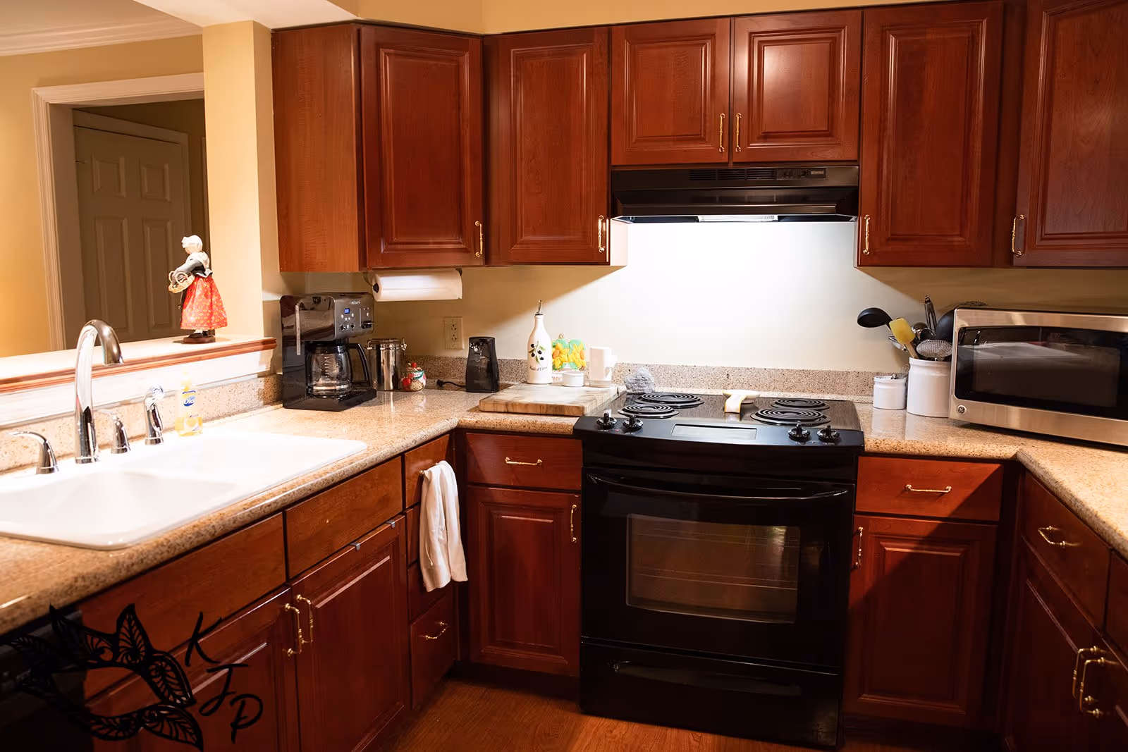 A kitchen with wooden cabinets, a black electric stove with oven, a microwave, a coffee maker, a white sink with a silver faucet, and various kitchen utensils on the countertop. There is a small decorative figurine on the counter near the sink.