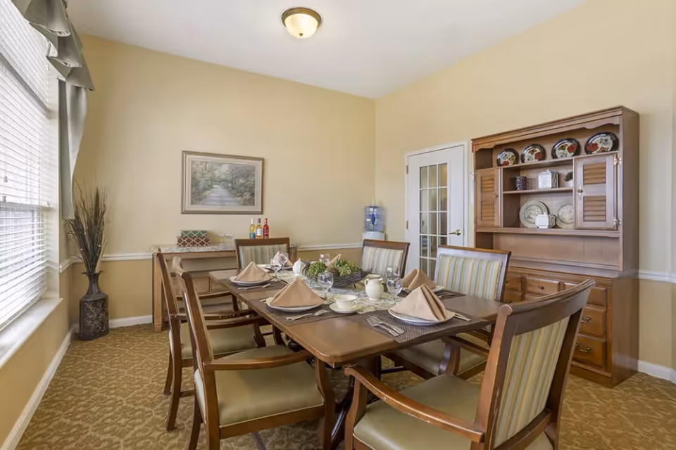 A dining room with a wooden table set for six people with beige napkins, plates, cups, and glasses. The room has beige walls, a window with blinds and curtains on the left, a wooden hutch with decorative plates on the right, and a small table with bottles and a water dispenser in the back corner.