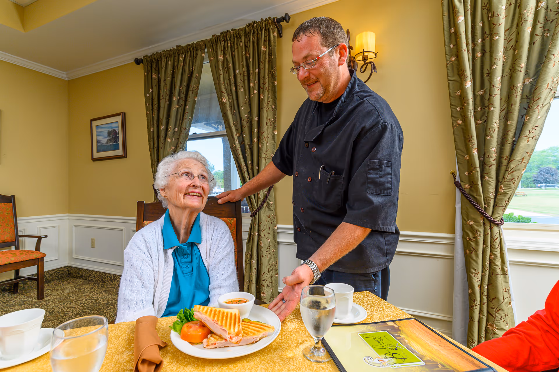 An elderly woman sits at a dining table smiling as a male server stands beside her presenting a plate with a sandwich in a dining room.