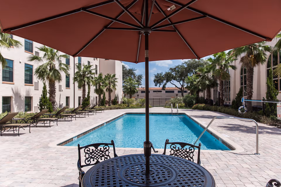 Outdoor swimming pool area at Lake Gibson Village with a round metal table and chairs under a large red umbrella in the foreground. The pool is surrounded by lounge chairs and palm trees, with a multi-story building in the background under a partly cloudy sky.