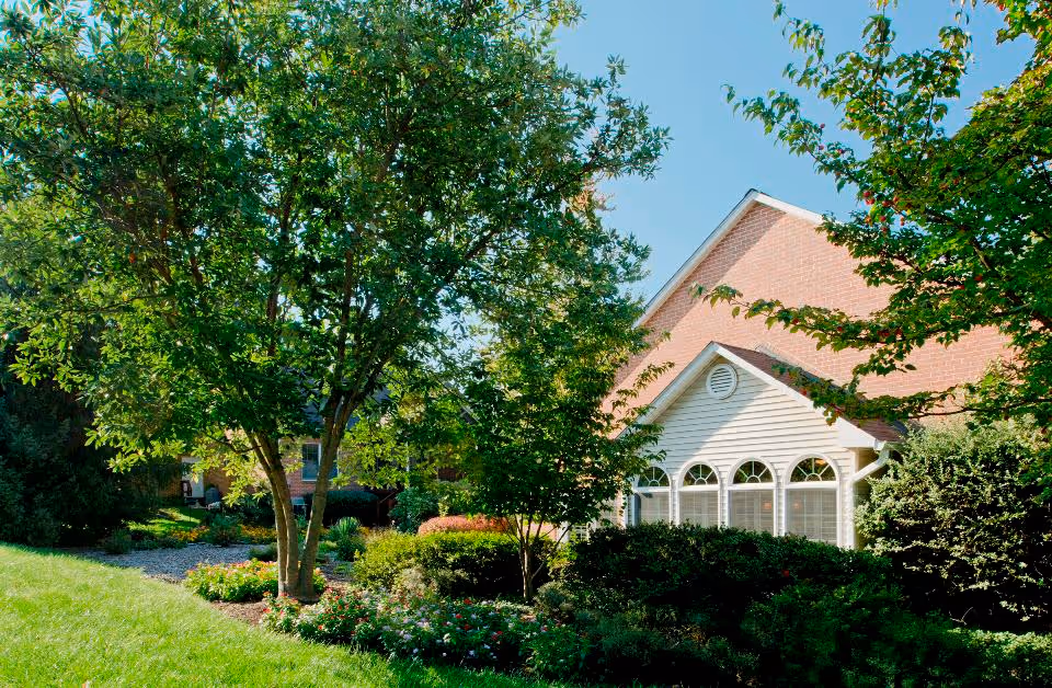 A sunny outdoor garden area with green grass, various trees, bushes, and flowering plants in front of a brick building with white siding and arched windows.