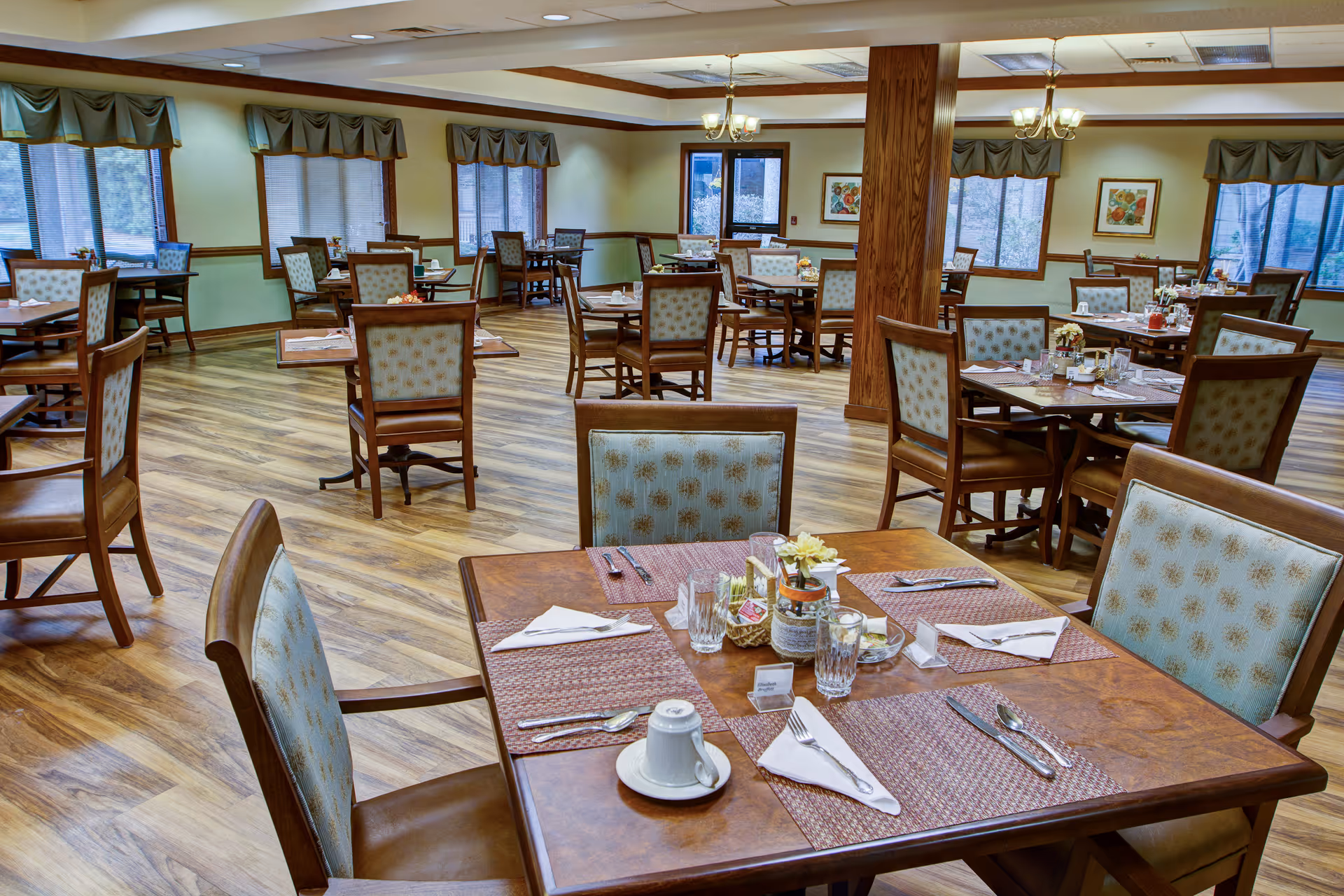 A spacious dining room with multiple wooden tables and chairs arranged neatly. Each table is set with placemats, utensils, napkins, glasses, and small decorative centerpieces. The room has large windows with green valances, wood trim, and light-colored walls. The floor is covered with wood-patterned flooring, and there are ceiling lights and chandeliers providing illumination.